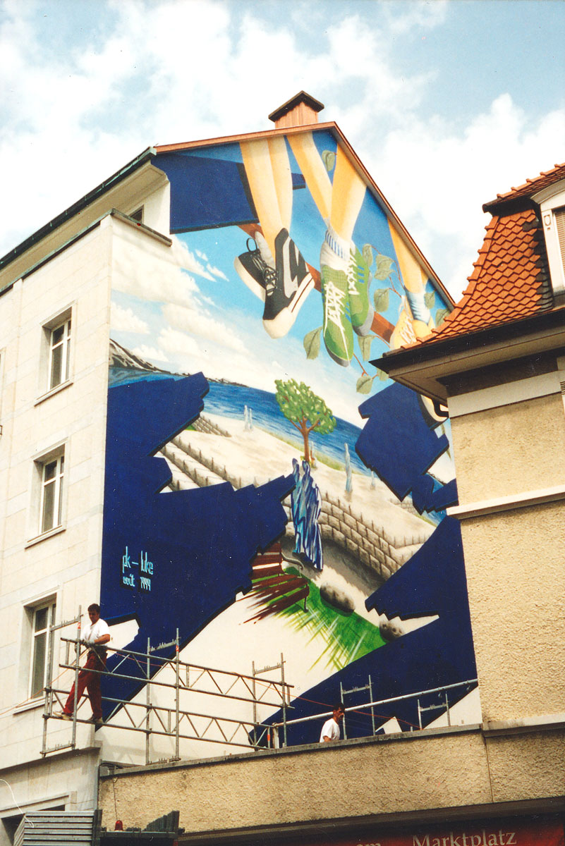 Wandbild auf Hausfassade mit überdimensionalen Beinen von Jugendlichen, die vom Dachfirst herunterbaumeln