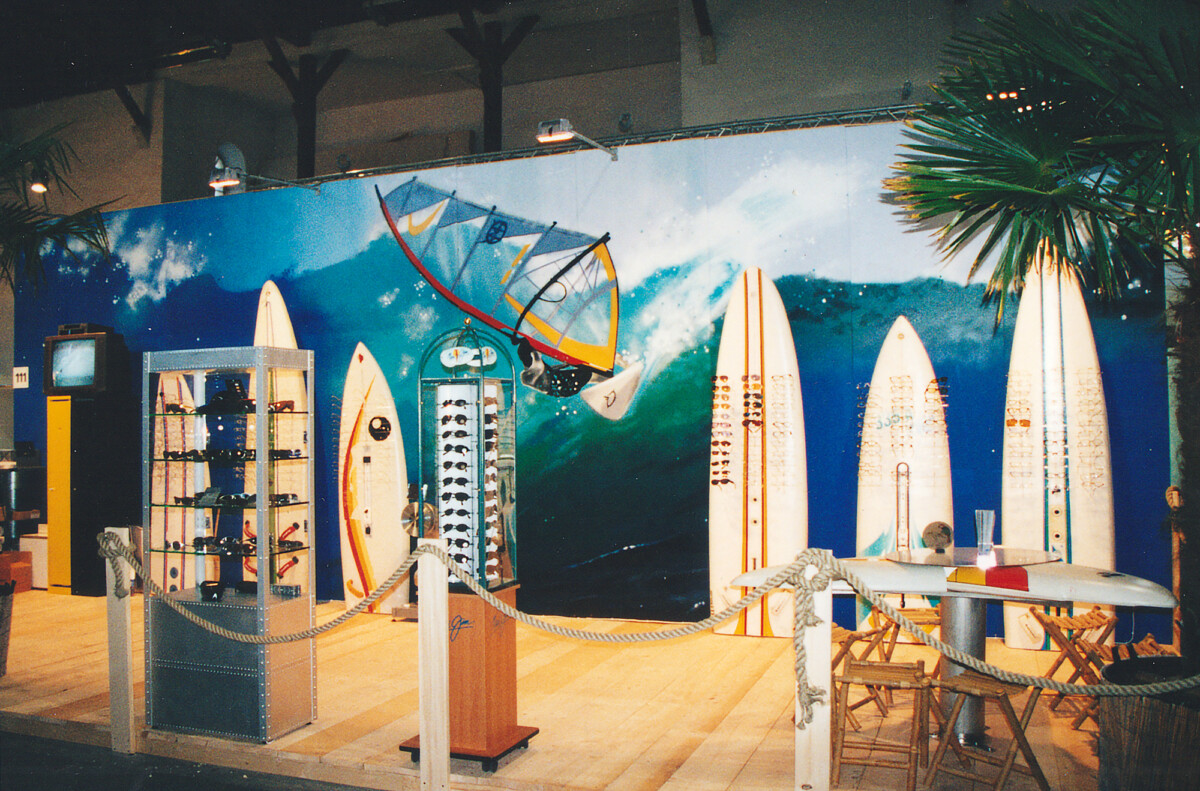 Gesprayter Windsurfer auf einer Welle, davor stehen Surfboards und Brillen